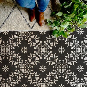 May include: A black and white patterned floor stencil with a floral design. The stencil is on a dark surface, with a person wearing blue jeans and brown boots standing above it. A beige rug and green plant are also visible.