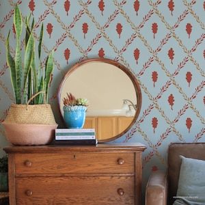 May include: A wooden dresser with a round mirror, a blue patterned wallpaper, and a potted snake plant in a woven basket. A small blue pot with succulents sits on top of a stack of books. The wallpaper has a light blue background with a repeating pattern of red oak leaves and vines.