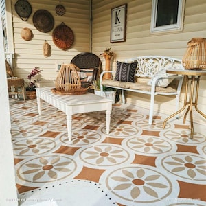 May include: A cozy outdoor living space with a white bench, a small round table, and a white coffee table. The floor features a repeating pattern of brown and beige floral designs. Woven baskets and a wooden lantern add to the rustic decor.
