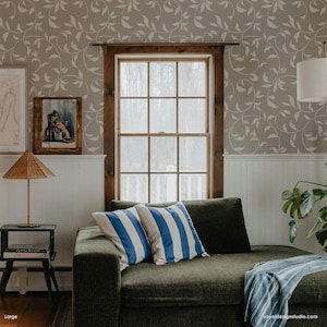 May include: A green couch with two blue and white striped pillows and a blue and white throw blanket. The couch is in front of a window with a view of trees. The wall behind the couch is covered in a beige wallpaper with a white vine pattern.