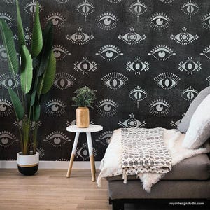 May include: A black and white eye pattern stencil on a black wall. The stencil is used to create a repeating pattern of eyes. A white side table with a gold vase and a plant sits in front of the wall. A gray couch with a white throw blanket is in the foreground.