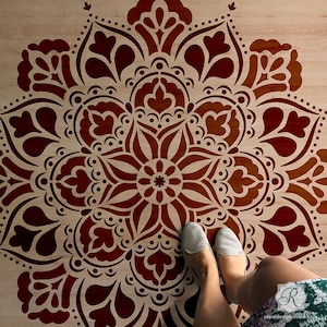 May include: A large, intricate mandala stencil with a brown background and a red design. The stencil is placed on a wooden floor and a person's feet are visible in the bottom of the image.