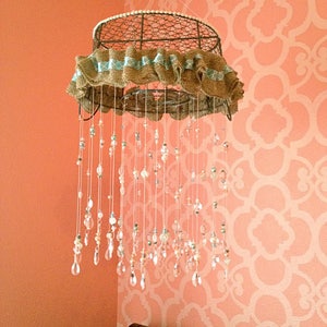 May include: A wire basket with a burlap and blue ribbon trim is hanging from the ceiling. It has strings of beads and crystal drops hanging from it. The basket is hanging in a room with a peach-colored wall and a white stencil pattern.