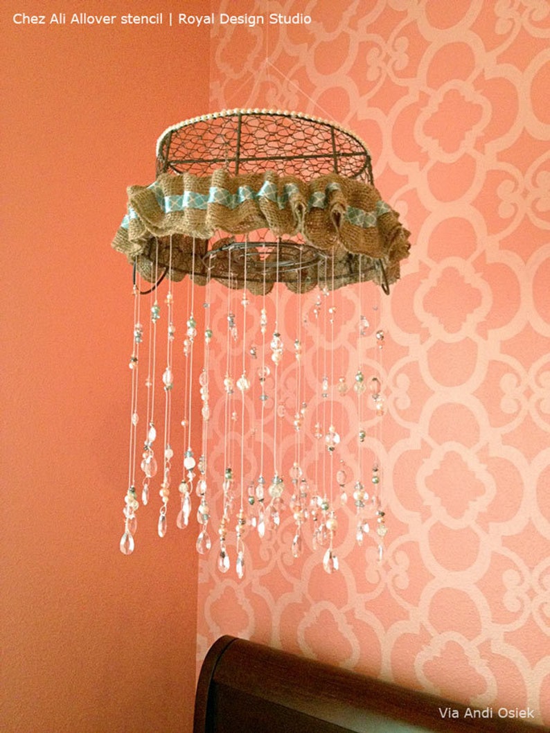 May include: A wire basket with a burlap and blue ribbon trim is hanging from the ceiling. It has strings of beads and crystal drops hanging from it. The basket is hanging in a room with a peach-colored wall and a white stencil pattern.