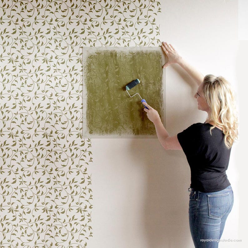 May include: A person using a paint roller to apply green paint to a wall stencil. The stencil is a leafy pattern and is being applied to a white wall. The wall behind the stencil is already painted with a green leafy pattern.