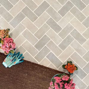 May include: A close-up of a herringbone pattern tile floor with a brown doormat in the foreground. There are three potted plants with pink and orange flowers on the floor.