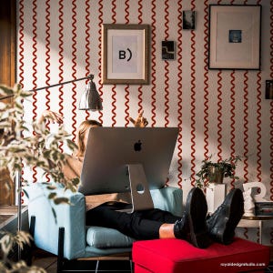 May include: A person is seated in a light blue armchair with their feet resting on a red ottoman, working on a large computer. The background features a white wall with red wavy lines and framed artwork. A silver desk lamp is positioned above the computer.