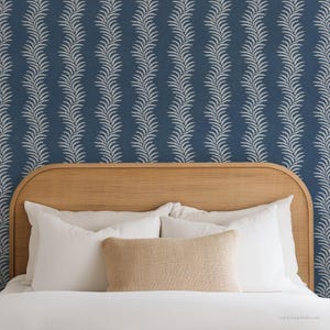 May include: A bedroom scene featuring a bed with white bedding, pillows, and a tan lumbar pillow. The headboard is made of light-colored wood. The wall behind the bed is covered in a blue wallpaper with a white leaf pattern.