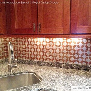 May include: A kitchen backsplash with a white and orange geometric pattern. The backsplash is made using a stencil and is illuminated by under-cabinet lighting. A stainless steel sink and faucet are in the foreground.