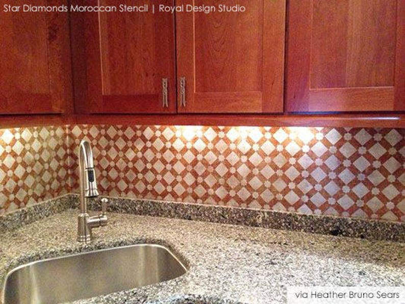 May include: A kitchen backsplash with a white and orange geometric pattern. The backsplash is made using a stencil and is illuminated by under-cabinet lighting. A stainless steel sink and faucet are in the foreground.