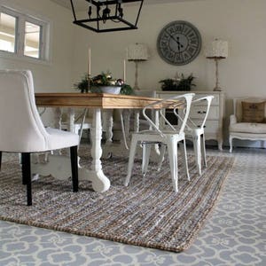 May include: A dining room with a wooden table and white chairs. The floor is painted with a gray and white Moroccan stencil pattern. A rug with a woven texture is on top of the painted floor. The room has a white ceiling with a black chandelier hanging from it. There is a clock on the wall with Roman numerals.