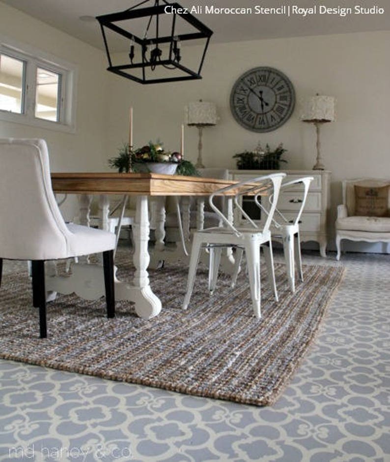 May include: A dining room with a wooden table and white chairs. The floor is painted with a gray and white Moroccan stencil pattern. A rug with a woven texture is on top of the painted floor. The room has a white ceiling with a black chandelier hanging from it. There is a clock on the wall with Roman numerals.