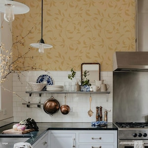 May include: A kitchen with white subway tile backsplash and a stainless steel range hood. The walls are covered in a yellow wallpaper with a vine pattern. There are two pendant lights hanging over the counter. A shelf above the counter holds a variety of kitchenware, including a set of copper pots, a glass pitcher, and a set of salt and pepper shakers.