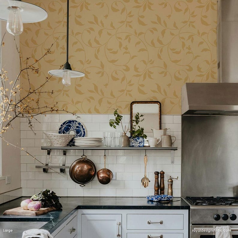 May include: A kitchen with white subway tile backsplash and a stainless steel range hood. The walls are covered in a yellow wallpaper with a vine pattern. There are two pendant lights hanging over the counter. A shelf above the counter holds a variety of kitchenware, including a set of copper pots, a glass pitcher, and a set of salt and pepper shakers.