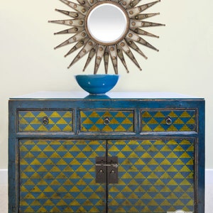 May include: A blue painted wooden cabinet with three drawers and two doors. The cabinet has a geometric pattern of yellow triangles on the front. A blue bowl sits on top of the cabinet, and a gold sunburst mirror hangs on the wall behind it.