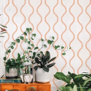 May include: A white wall with a repeating orange leaf pattern serves as a backdrop for a collection of potted plants. The plants, in various sizes and shapes, sit on a wooden cabinet. A book titled "Botanica" is visible.