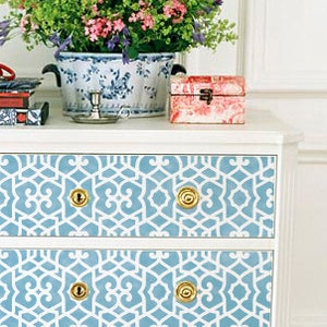 May include: White dresser with two drawers featuring a blue and white geometric pattern. The drawers have gold knobs. A blue and white ceramic planter with green plants and a small red box are on top of the dresser.