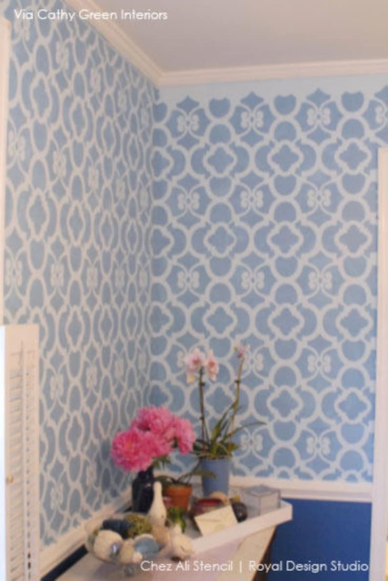 May include: A blue wall with a white stencil pattern. The wall is decorated with pink flowers in a pot. The text "Chez Ali Stencil | Royal Design Studio" is visible at the bottom of the image.