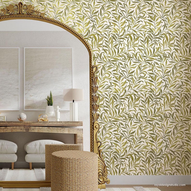 May include: A gold-framed mirror with an ornate design reflects a room with a wooden table, two white ottomans, and a woven stool. The wall behind the mirror is covered in a wallpaper with a repeating pattern of green leaves on a white background.