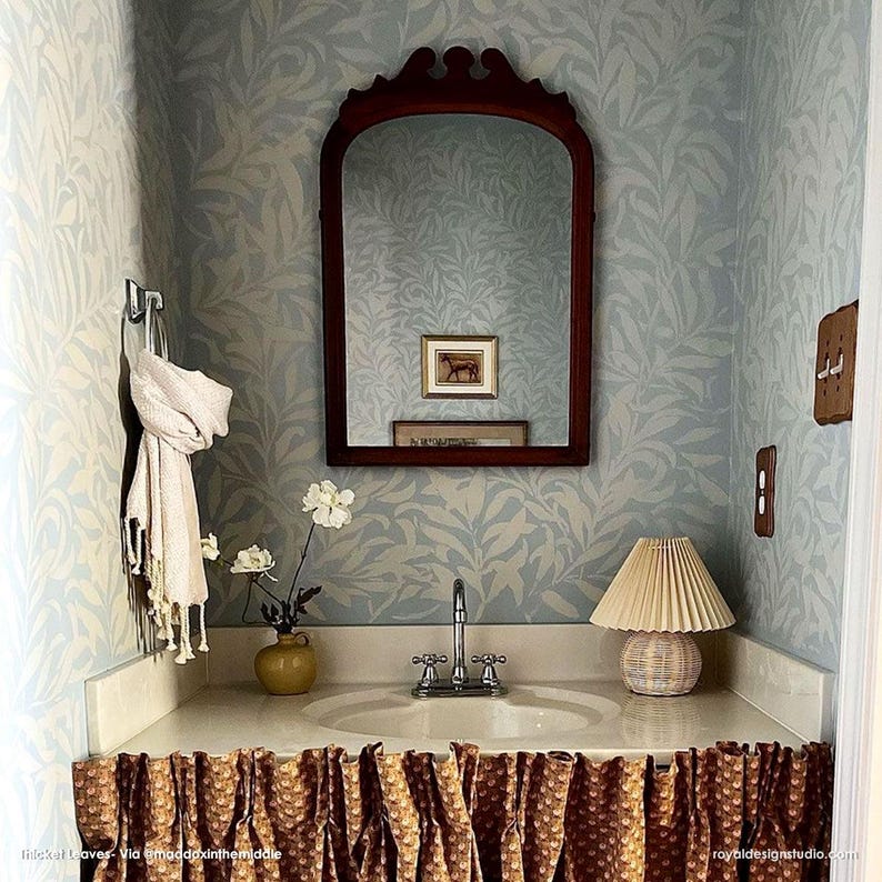 May include: A bathroom vanity with a brown patterned fabric skirt, a white porcelain sink, and a silver faucet. A white lamp with a pleated shade sits on the counter next to a small brown vase with white flowers. A mirror with a dark wood frame hangs above the sink. The walls are covered in a light blue wallpaper with a white leaf pattern.