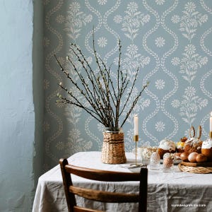 May include: A light blue wall with a white floral stencil pattern. A table with a white tablecloth is set with a wicker vase filled with willow branches. The table is decorated with Easter bread, eggs, and a wicker serving tray.