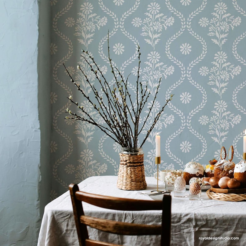 May include: A light blue wall with a white floral stencil pattern. A table with a white tablecloth is set with a wicker vase filled with willow branches. The table is decorated with Easter bread, eggs, and a wicker serving tray.