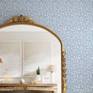 May include: A blue and white wallpaper stencil with a delicate vine pattern. The stencil is shown in a room with a gold mirror, two framed paintings, a wooden table, and a lamp.