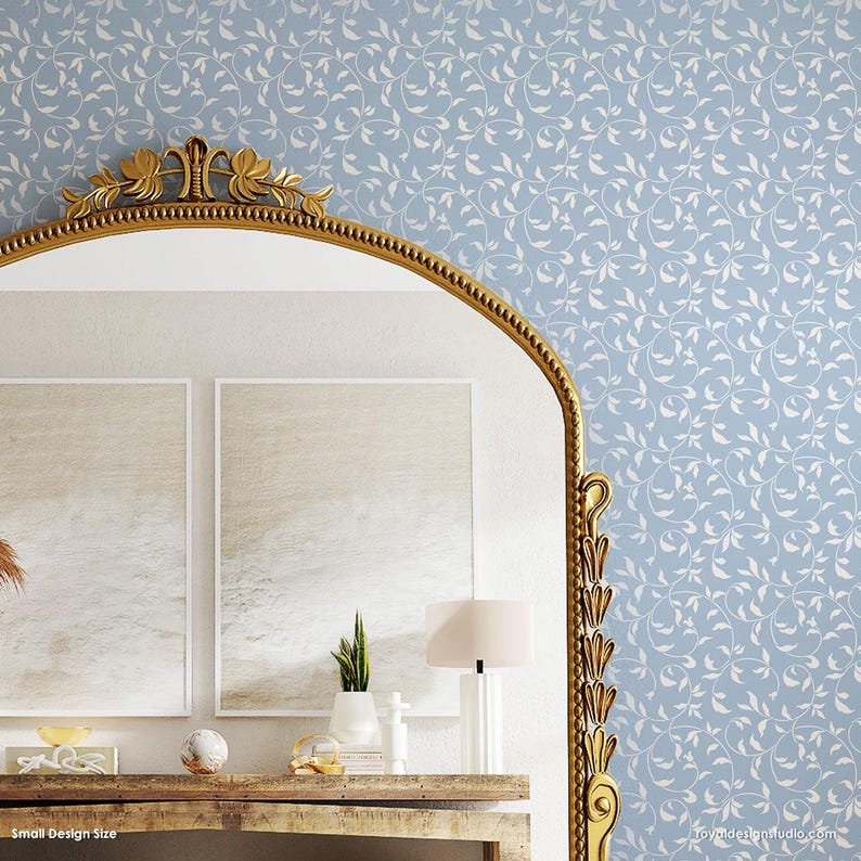 May include: A blue and white wallpaper stencil with a delicate vine pattern. The stencil is shown in a room with a gold mirror, two framed paintings, a wooden table, and a lamp.