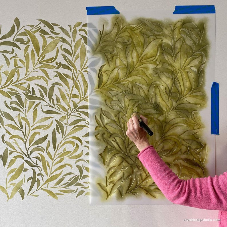 May include: A person using a stencil and a brush to paint a green leafy pattern on a wall. The stencil is a repeating pattern of leaves and branches.