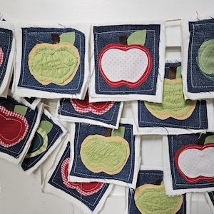 May include: A handmade apple-themed banner. Each square features a denim background with a white border and a fabric apple applique. The apples are red, green, and yellow with green leaves. The banner is strung on a natural twine.