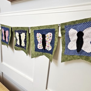 May include: A decorative butterfly banner with six fabric flags. Each flag displays a butterfly design in shades of pink, white, and floral patterns, set against a blue or patterned backdrop, and framed by green fabric.