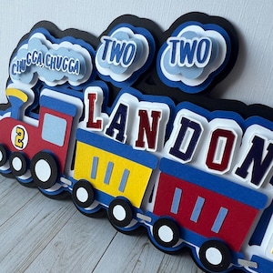 May include: A colourful paper train cake topper in red, yellow, and blue. The train cars spell out "LANDON" and include the number "2" and the words "CHUGGA CHUGGA" and "TWO". The topper is designed for a birthday celebration.
