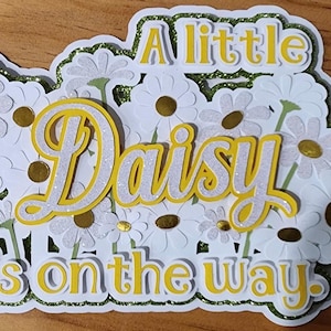 May include: A layered paper craft featuring white daisies with gold centers and green glitter accents. The words "A little Daisy is on the way" are in yellow and white, suggesting a baby shower or announcement theme.
