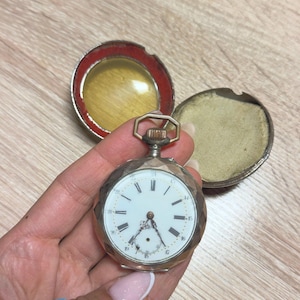 Puede incluir: Un reloj de bolsillo plateado con esfera blanca y números negros. El reloj tiene un diseño geométrico en la caja y está abierto, revelando el mecanismo interno. El reloj se sostiene en una mano, con un anillo de cuentas en el dedo.