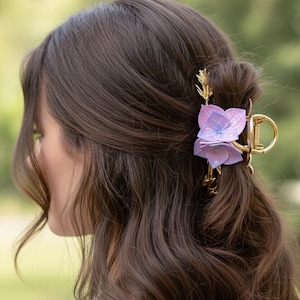 May include: A gold-coloured hair clip adorns brown hair. The clip features a cluster of purple and lavender flower accents. This decorative hair accessory is designed to hold the hair in place.