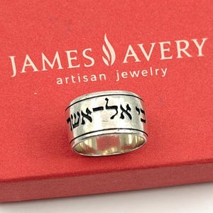 May include: A wide sterling silver band ring featuring engraved black Hebrew lettering around the exterior surface. The ring is positioned in front of a red box with the white text JAMES AVERY artisan jewelry printed on the top surface.