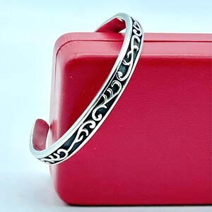 May include: A silver cuff bracelet featuring a continuous raised scrollwork pattern against a dark, recessed background. The metal has a polished finish on the edges and the intricate swirling design, creating a high-contrast look.