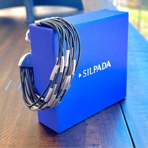 May include: A multi-strand black leather bracelet with silver-tone metal accents is displayed on a blue box. The box has the brand name "SILPADA" printed on it. The bracelet is a modern jewelry piece.