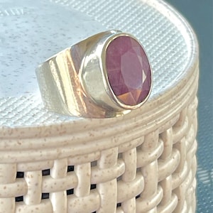 May include: A silver ring with a large, oval-cut ruby gemstone. The ruby is a deep red color and is set in a polished silver band. The ring has a classic, elegant design.