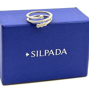 May include: A silver-toned bypass ring with a textured, beaded band and three small, clear round stones set at the ends. The ring rests on a rectangular, textured royal blue box with the white text SILPADA printed on the front face.