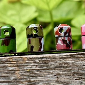 May include: Four small, cylindrical bottle stoppers with a camouflage pattern in green, brown, pink, and tan. Each stopper has a hole at the top. The stoppers are displayed on a wooden surface with a blurred green background.