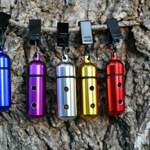 May include: A collection of colorful, cylindrical metal containers in blue, purple, silver, gold, and red, each with three circular holes. They are suspended from black clips against a textured brown background.