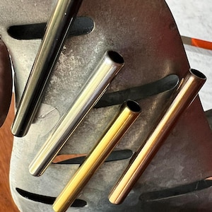 May include: An assortment of metal tubes in various colors, including black, silver, and gold. The tubes are cylindrical with a hollow interior and are arranged on a textured metal surface. The tubes are of similar length and diameter.