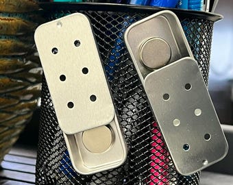 May include: Two open, rectangular, silver-colored metal containers with hinged lids. Each container holds a round, silver-colored magnet. The lids have several evenly spaced holes. The containers are positioned in front of a black wire mesh.