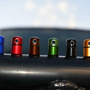 May include: A row of six cylindrical objects in various colors: blue, red, bronze, green, black, and gold. Each has a circular hole at the top. They are arranged on a dark, curved surface, with a blurred background.