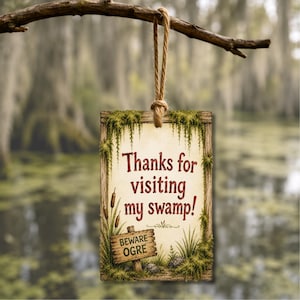 Pode incluir: Uma placa retangular suspensa por uma corda, com um centro creme contendo o texto "Thanks for visiting my swamp!" em vermelho, e uma placa de madeira menor abaixo com "BEWARE OGRE", cercada por ilustrações de musgo e taboas.