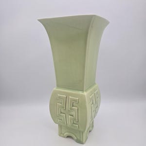 May include: A celadon green ceramic vase with a flared top and a square base. The vase features a geometric design on the sides and is approximately 12 inches tall. The vase has a glossy finish and a traditional aesthetic.