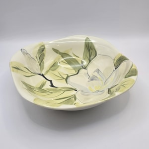 May include: Oval-shaped ceramic serving bowl with a hand-painted magnolia design. The bowl features a creamy white base with a floral pattern in shades of green, gray, and white. The bowl is approximately 12 inches in diameter.