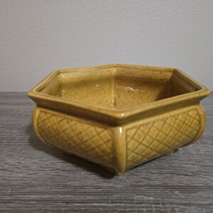 May include: A square, ceramic planter with a textured, diamond pattern on the sides. The planter is a warm, speckled yellow color. The top edge has a simple, smooth rim. The planter sits on a gray wooden surface.