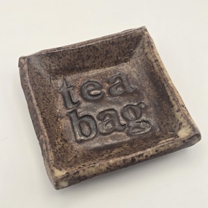 May include: A set of three ceramic mugs with a speckled, matte finish. The mugs are in shades of cream, light brown, and dark brown. Each mug has a simple, minimalist design and a rounded handle. The mugs are likely for coffee or tea.